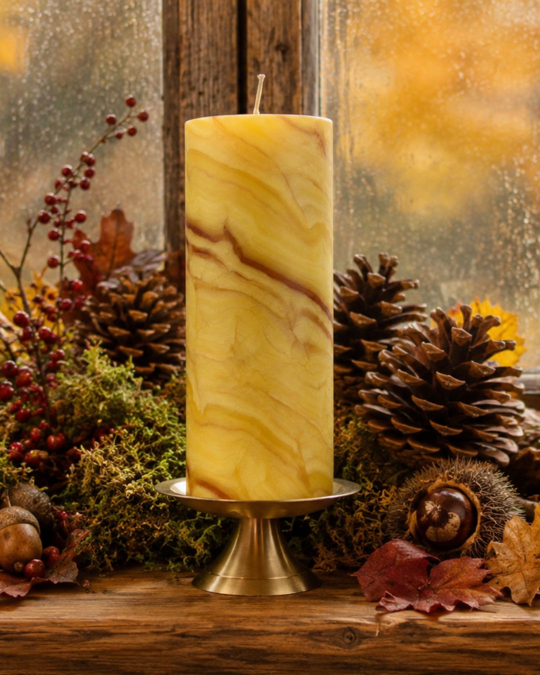 Handgeknetete Bienenwachskerze in Naturfarben vor einem Herbstfenster mit Moos, Kastanien, Tannenzapfen und roten Beeren.