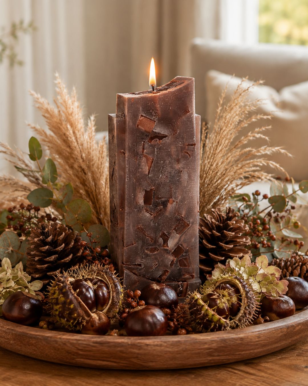 Dunkelbraune Designkerze mit Kastanien, Zapfen und Pampasgras auf einem rustikalen Holztablett im herbstlich dekorierten Wohnzimmer.